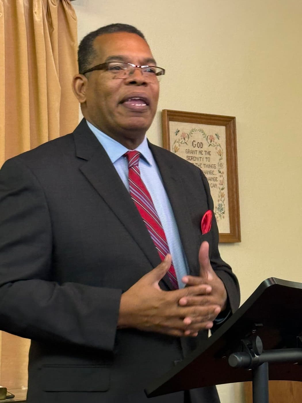 A pastor preaching from a pulpit with the Serenity Prayer framed on the wall behind him.